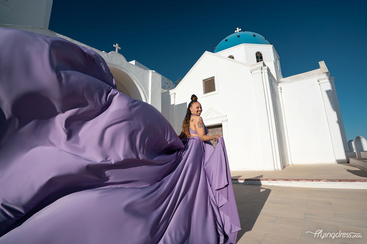 A lavender transformer dress swirls dramatically through Santorini's whitewashed alleyways with the iconic blue-domed church as a stunning backdrop.